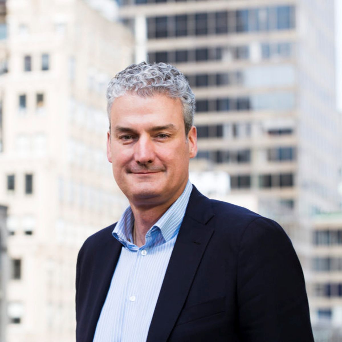 John MacPhee in navy blazer smiling outdoors with office buildings in background
